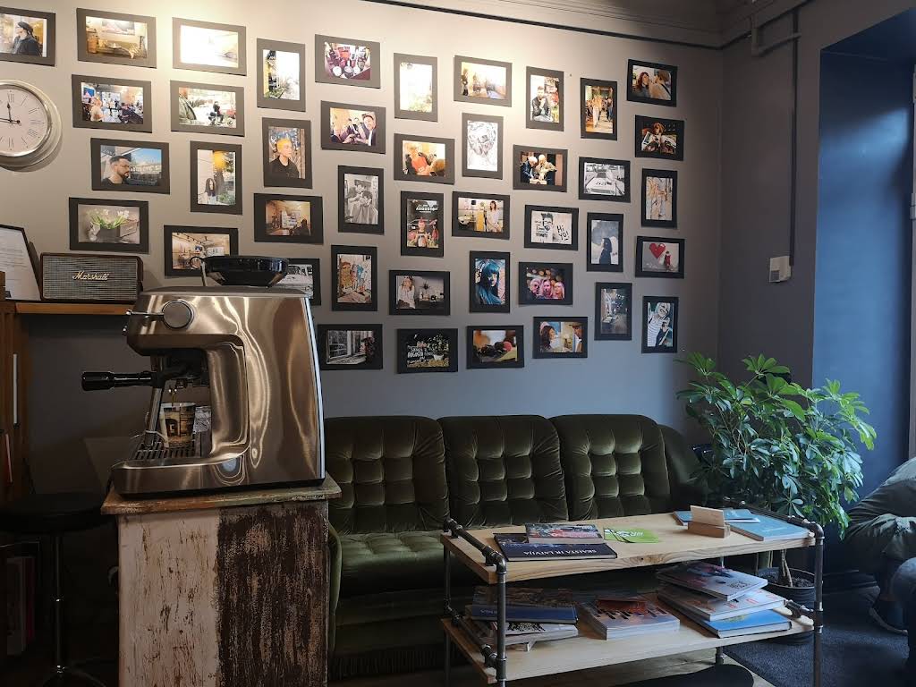 Salonа interjers — fotogrāfiju siena, espresso mašīna un zaļais samtainais dīvāns