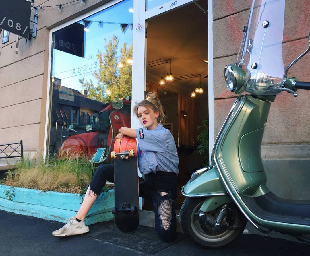 Jauna sieviete ar skrituļdēli pie salonа — zaļais Vespa motorollers blakus un sarkanais Vespa redzams logā